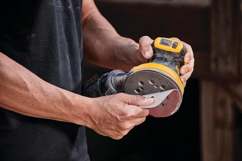 Una persona sostiene una lijadora orbital inalámbrica DEWALT, modelo DCW210B, colocando un disco de lija en la herramienta.