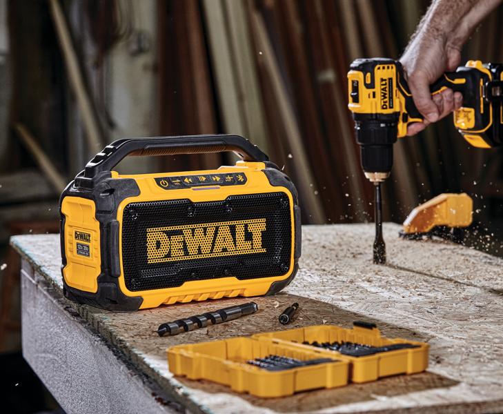 Un altavoz Bluetooth portátil DEWALT está colocado sobre un banco de trabajo junto a un taladro inalámbrico DEWALT en uso, brocas y una caja organizadora amarilla, todo en un taller.