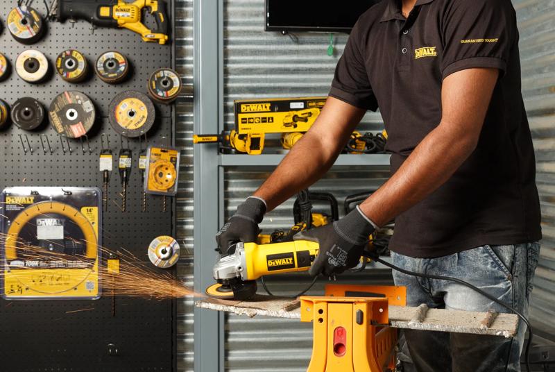 Una persona con guantes utiliza una amoladora angular Dewalt DW803 para cortar una pieza de metal, y se ven chispas. Herramientas eléctricas Dewalt y discos para esmerilado están exhibidos en una pared y una mesa de trabajo en el fondo.